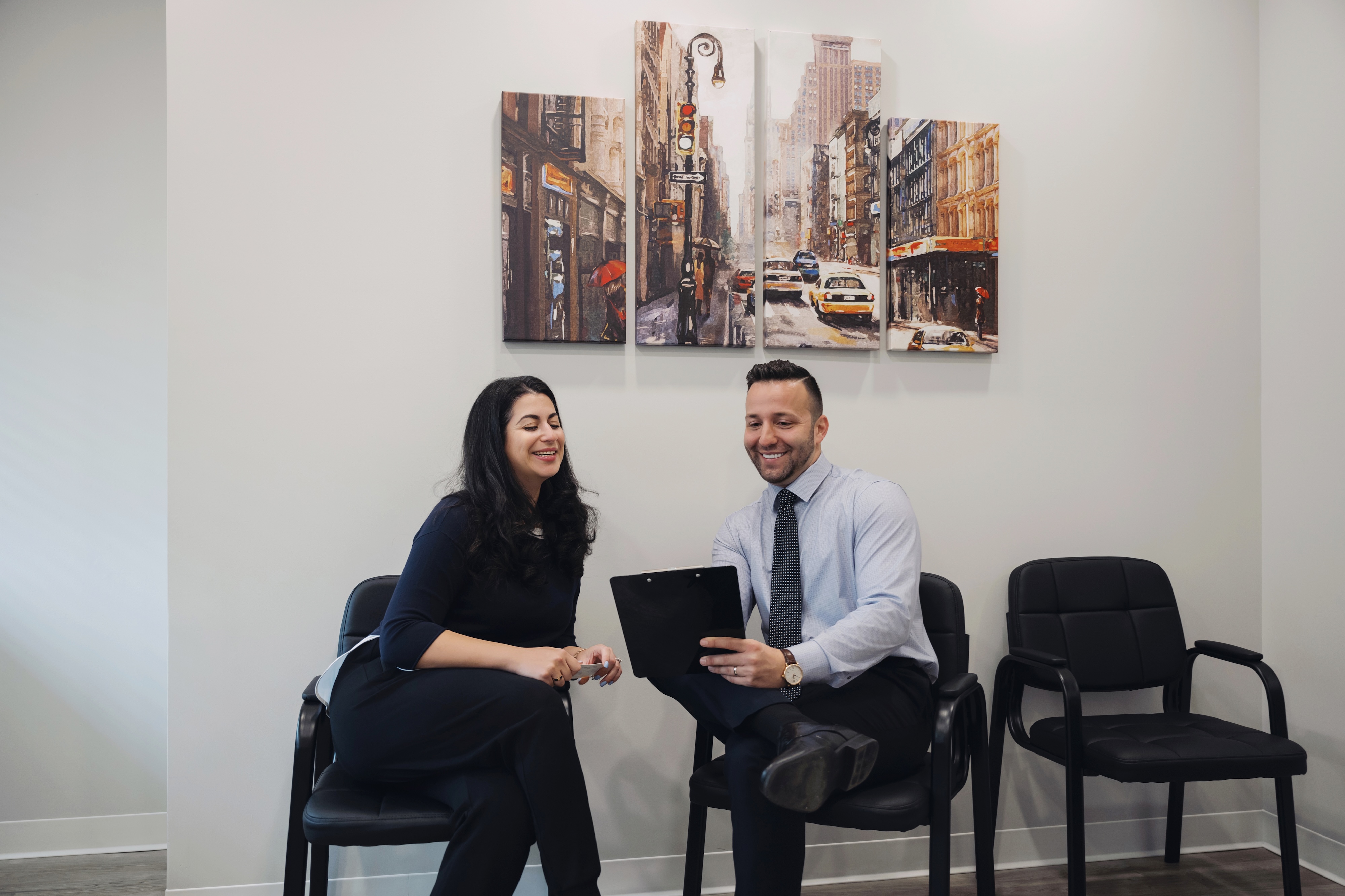 The dentist and the dental patient are both sitting in chairs next to each other in the waiting area while the dentist shows a clipboard full of information 