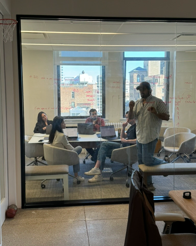 Five people in a glass-walled office conference room engaged in a meeting with laptops open and red writing on the glass wall.