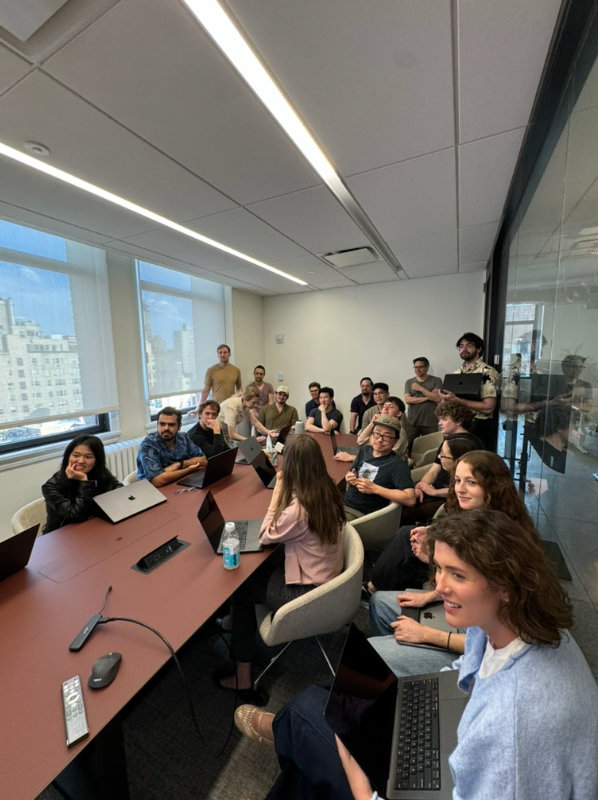 Group of diverse young adults sitting and standing around a conference table with laptops in a bright office meeting room.
