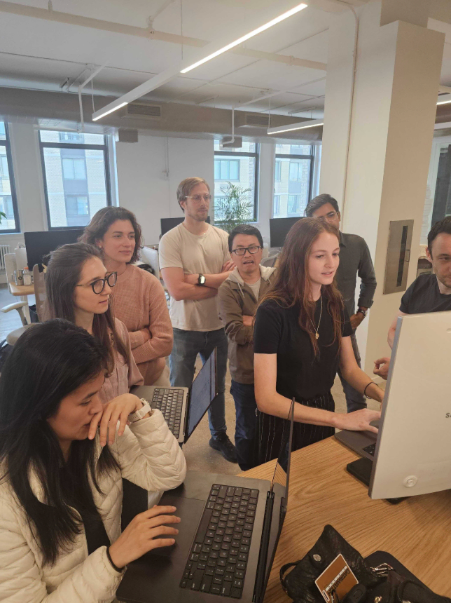 A group of eight colleagues attentively gathered around a computer screen in a modern office setting.