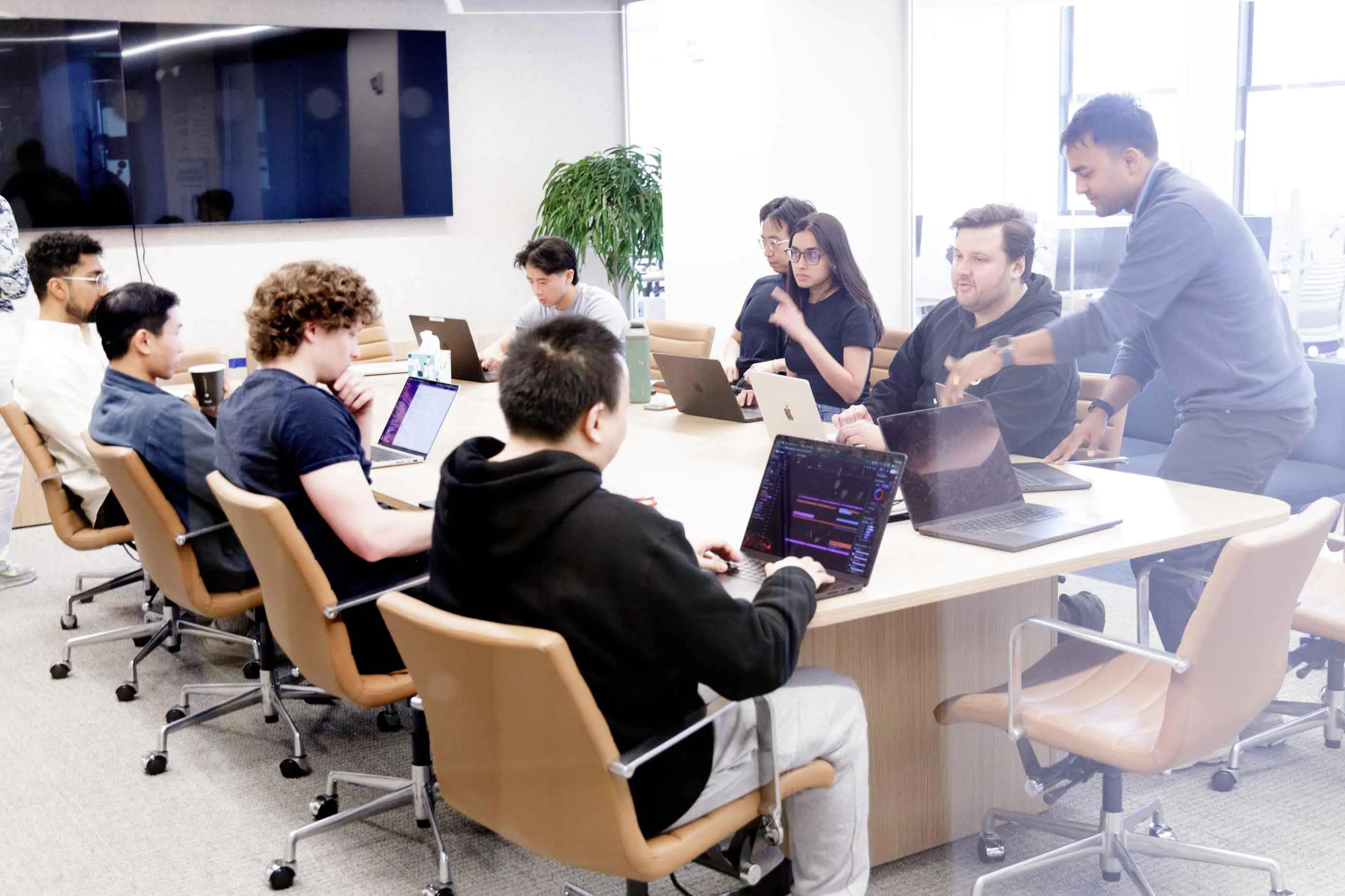 A diverse group of people collaborating around a conference table with laptops in a bright office.