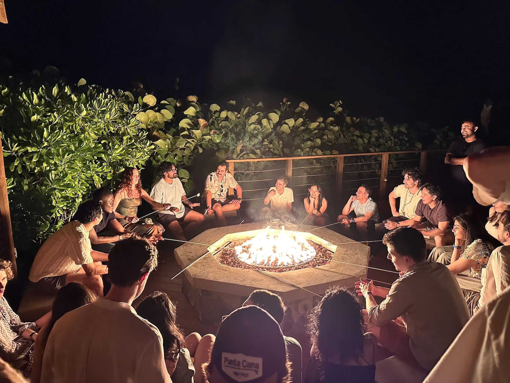 A large group of around twenty people gather around an octagonal stone fire pit at night, many holding long roasting sticks with marshmallows over the glowing flames. They're surrounded by tropical greenery and warm string lights, all laughing and engaged in conversation.