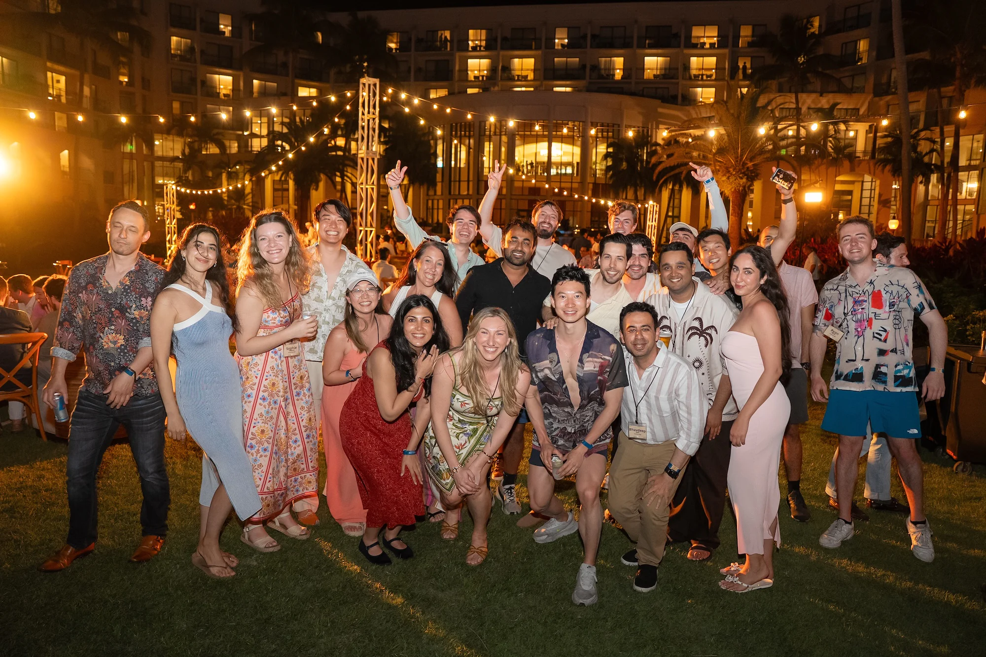 A large group of roughly twenty-five people pose together outdoors at night, dressed in colorful summer attire. String lights and palm trees frame a large hotel building in the background, and several people in the back are raising their hands and cheering.
