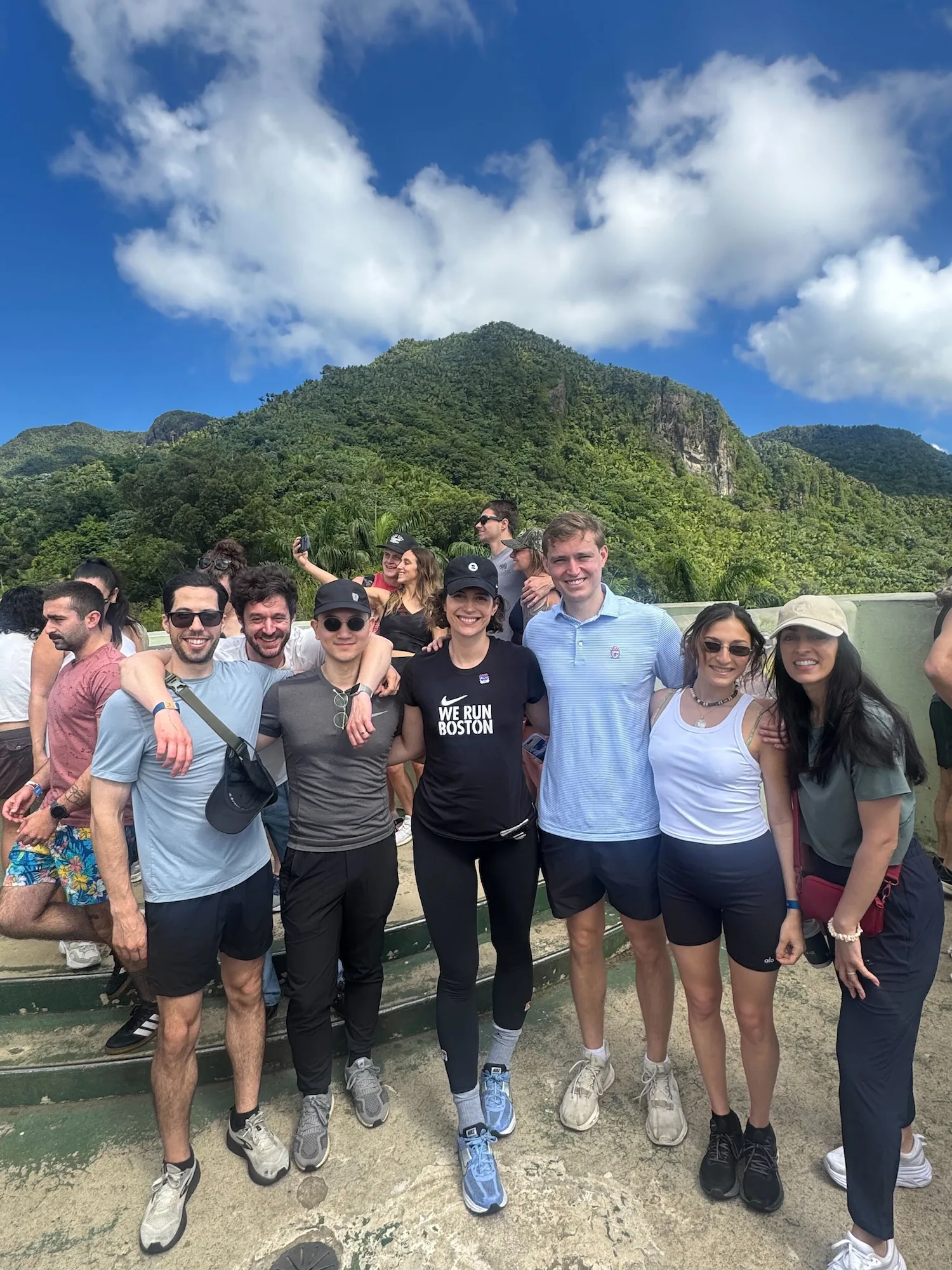 A group of six coworkers pose together smiling at a scenic overlook, with a lush green mountain peak rising dramatically behind them under a bright blue sky with clouds. The group is dressed in athletic wear, with one person wearing a black "We Run Boston" Nike shirt.