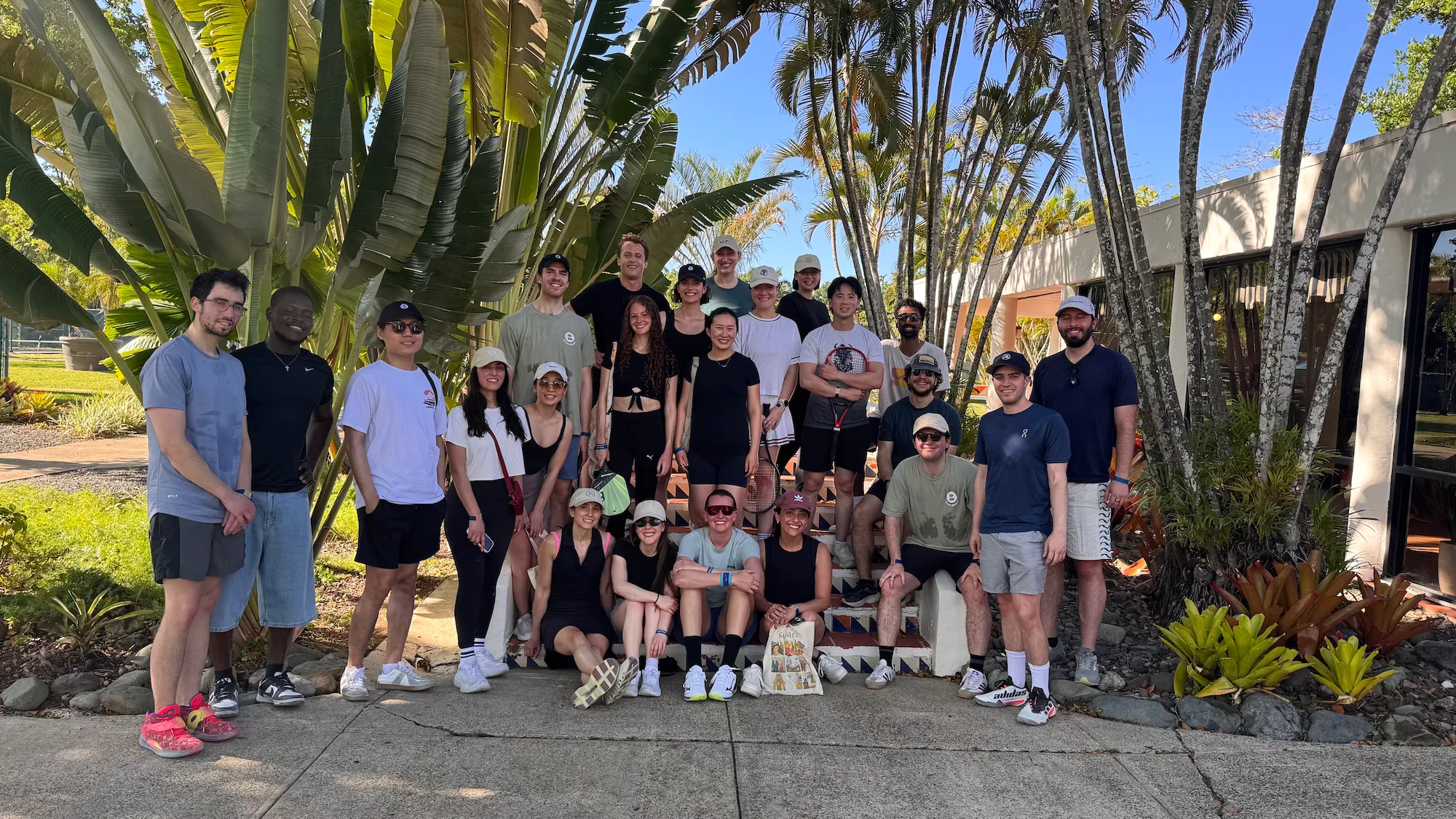 A group of approximately twenty-five people pose together on and around a set of decorative tiled steps, surrounded by lush tropical landscaping including large banana leaves and tall palm trees under a clear blue sky. The group is dressed in casual summer and athletic wear, with several people holding tennis rackets, suggesting a post-activity group photo at a resort or club.
