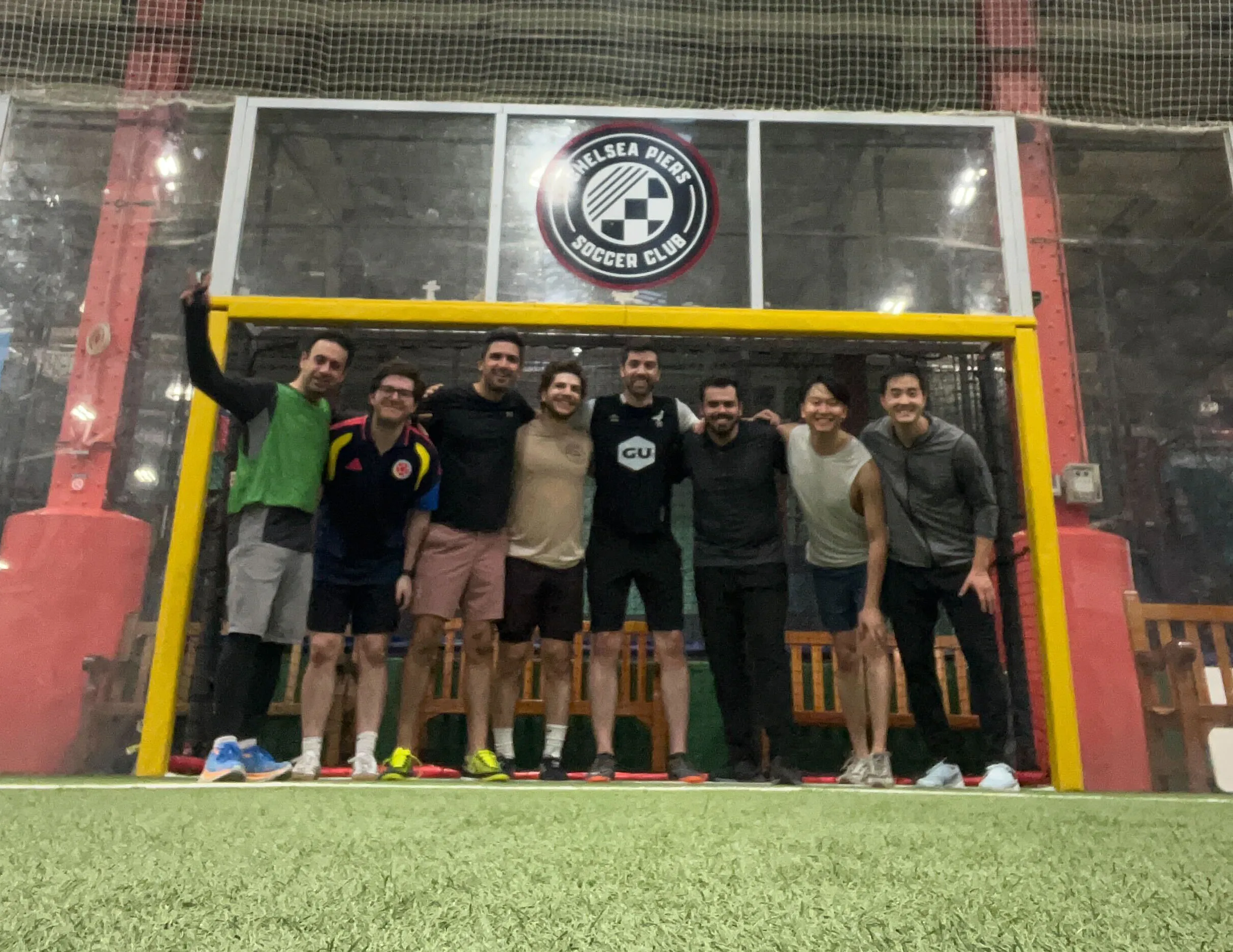A group of eight men posing together in front of a yellow soccer goal at Chelsea Piers Soccer Club indoor facility.