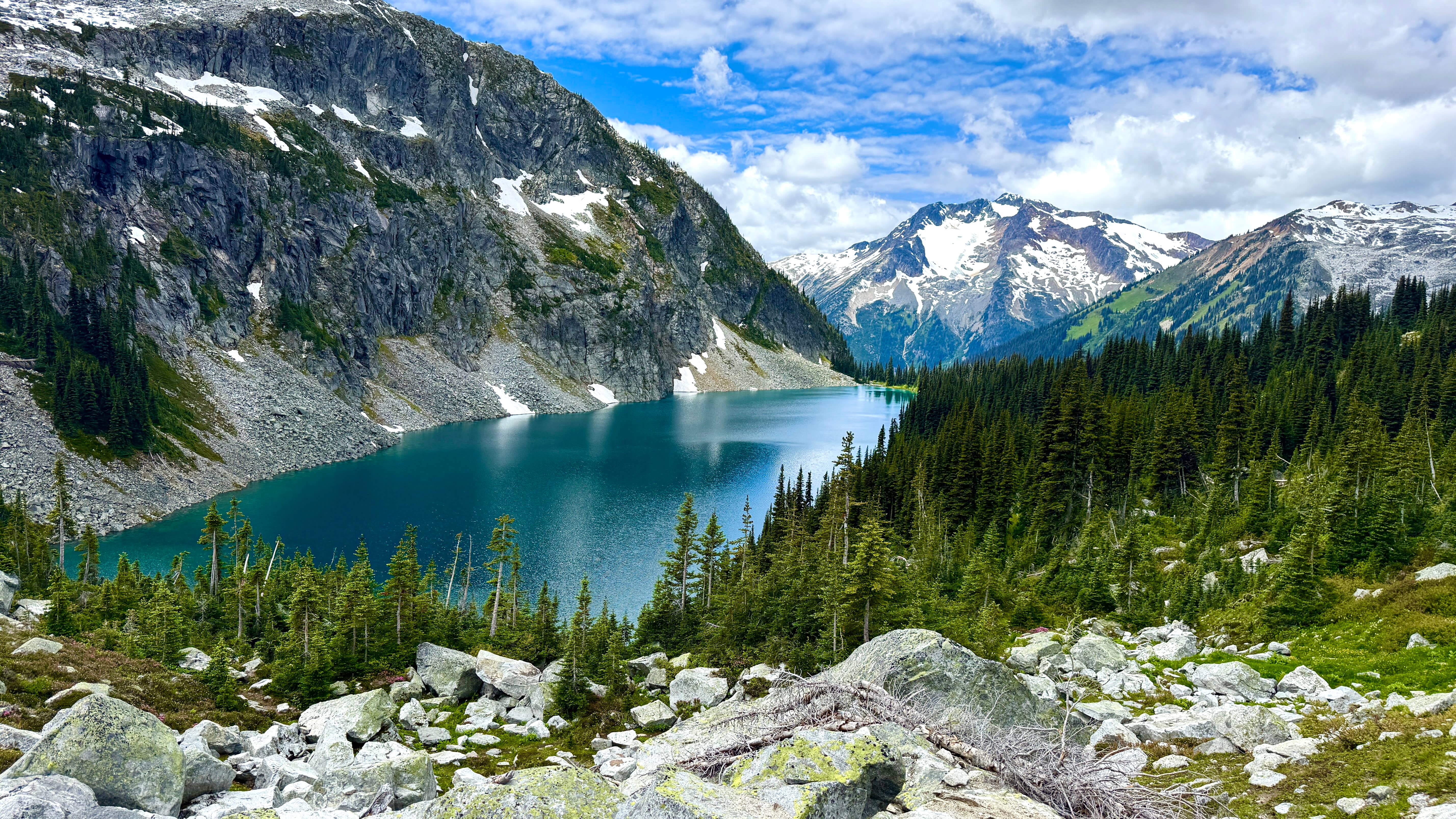 Scenic view of Rohr Lake in British Columbia, featuring vibrant turquoise water surrounded by dense evergreen forests, rocky shores, and steep mountain slopes under a clear blue sky