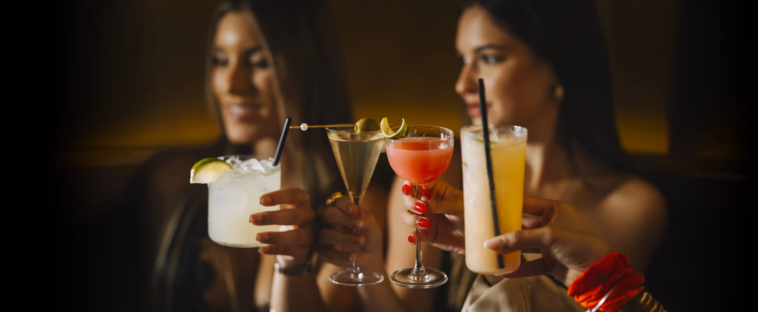 Four women holding different cocktails including a margarita, martini, pink cocktail, and a tall yellow drink with straws in a dimly lit bar.