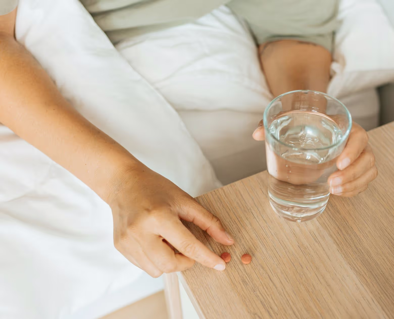 A person picking up a prescription tablet from a nightstand while holding a glass of water.