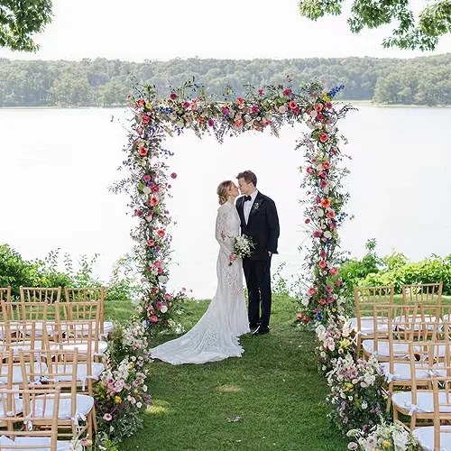 A bridge and groom under an arch of flowers with a lake in the background.