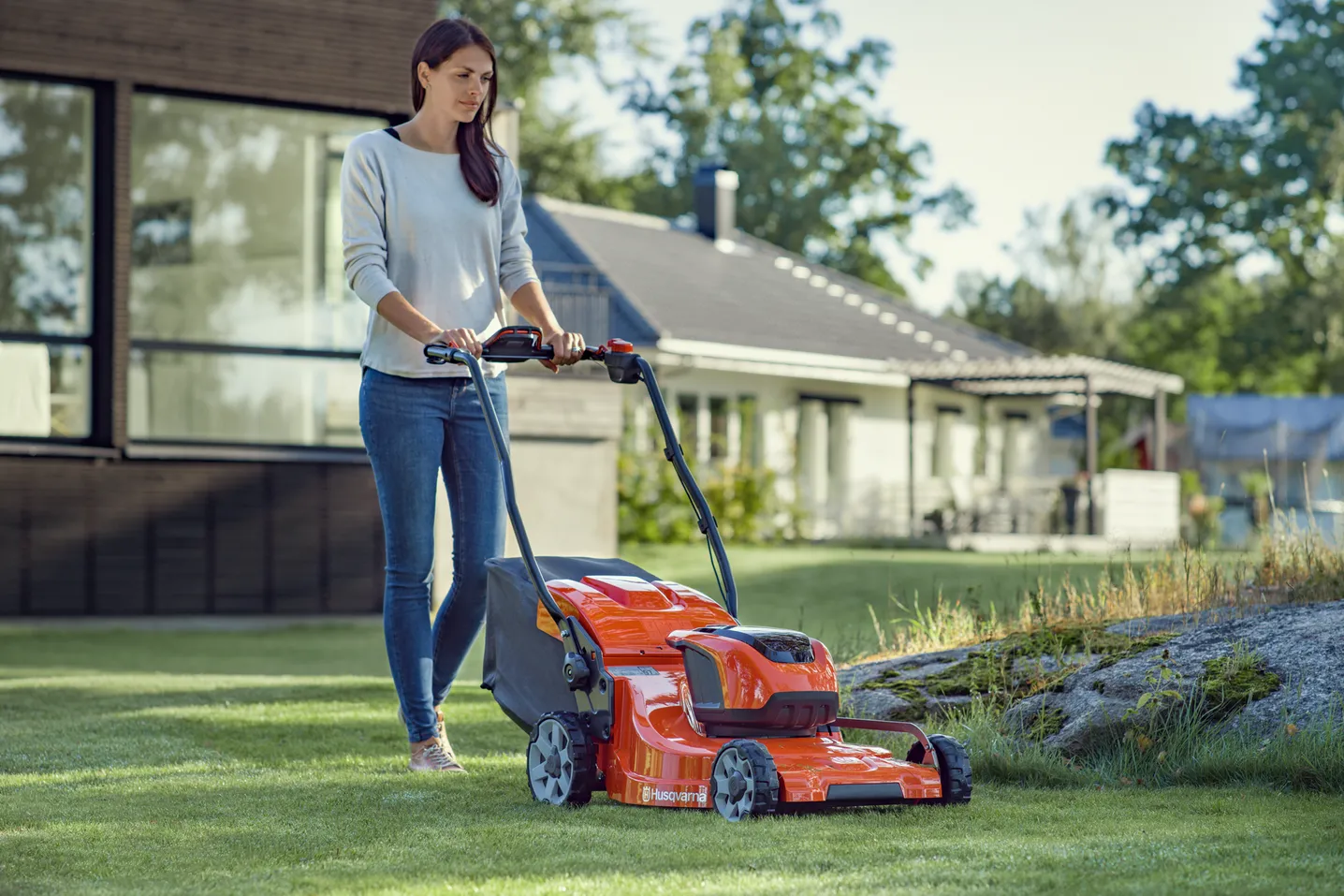 Woman mowing a green lawn with an orange Husqvarna lawn mower in a residential backyard.