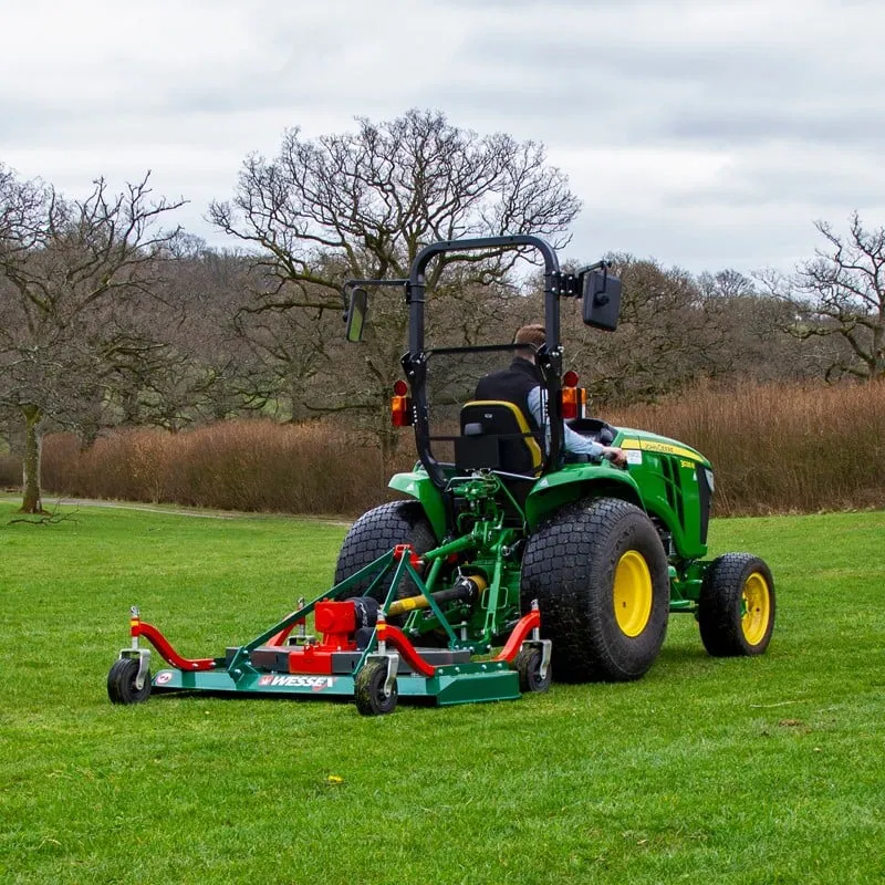 Person driving a green and yellow tractor with a large mower attachment cutting green grass on a cloudy day.