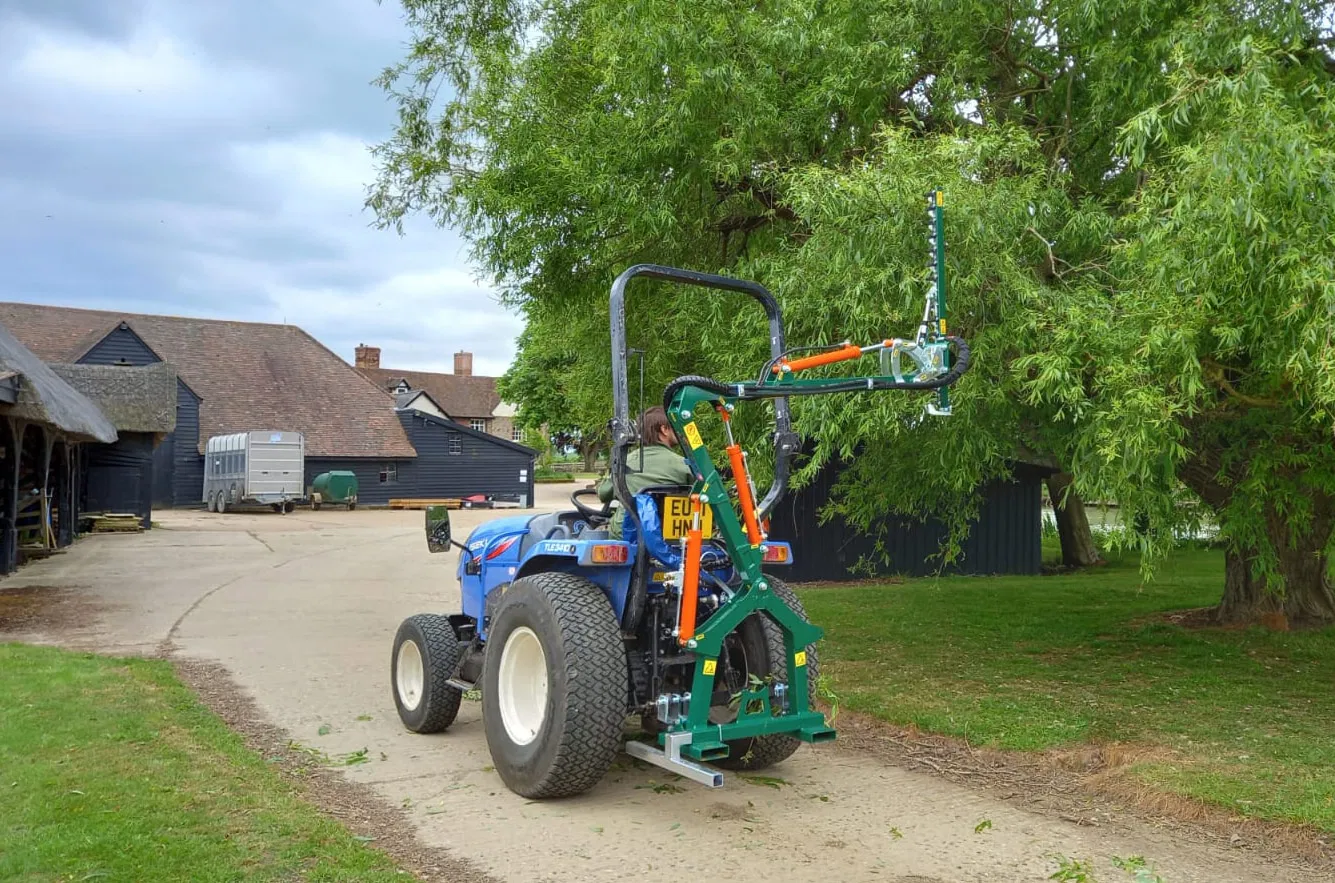 Blue compact tractor equipped with a green hydraulic tree pruner trimming low branches in a rural farmyard.