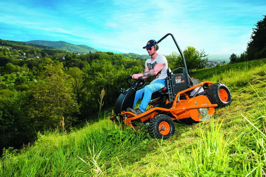 Man wearing sunglasses and ear protection driving an orange all-terrain lawn mower on a steep grassy hillside with forested hills in the background.