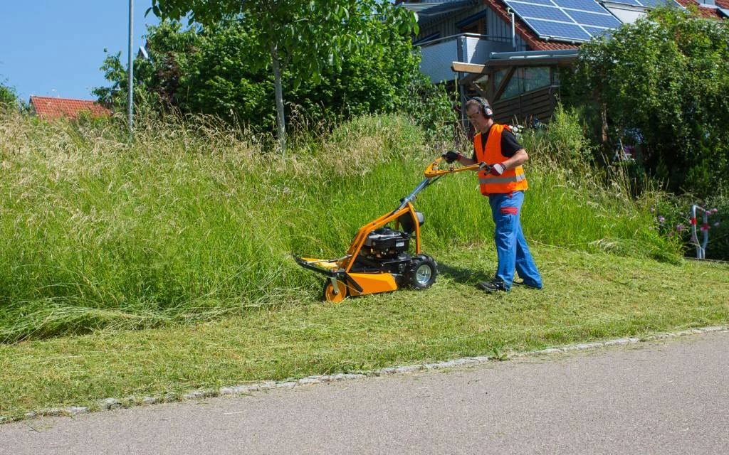 Man wearing orange safety vest and headphones mowing tall grass with a push mower near a residential area with solar panels.