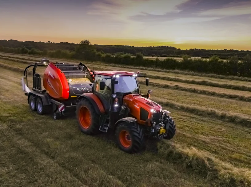 Orange tractor pulling a hay baler across a field at sunset.