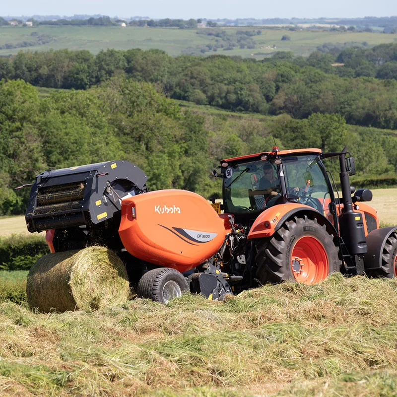Orange Kubota BF3500 tractor baling hay in a grassy field with green trees and hills in the background.