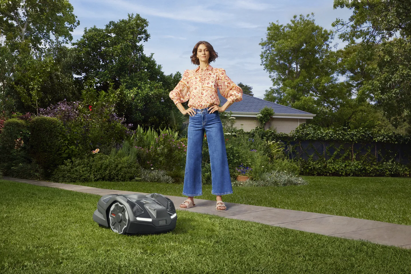 Woman standing on a garden path with hands on hips next to a robotic lawn mower on green grass.