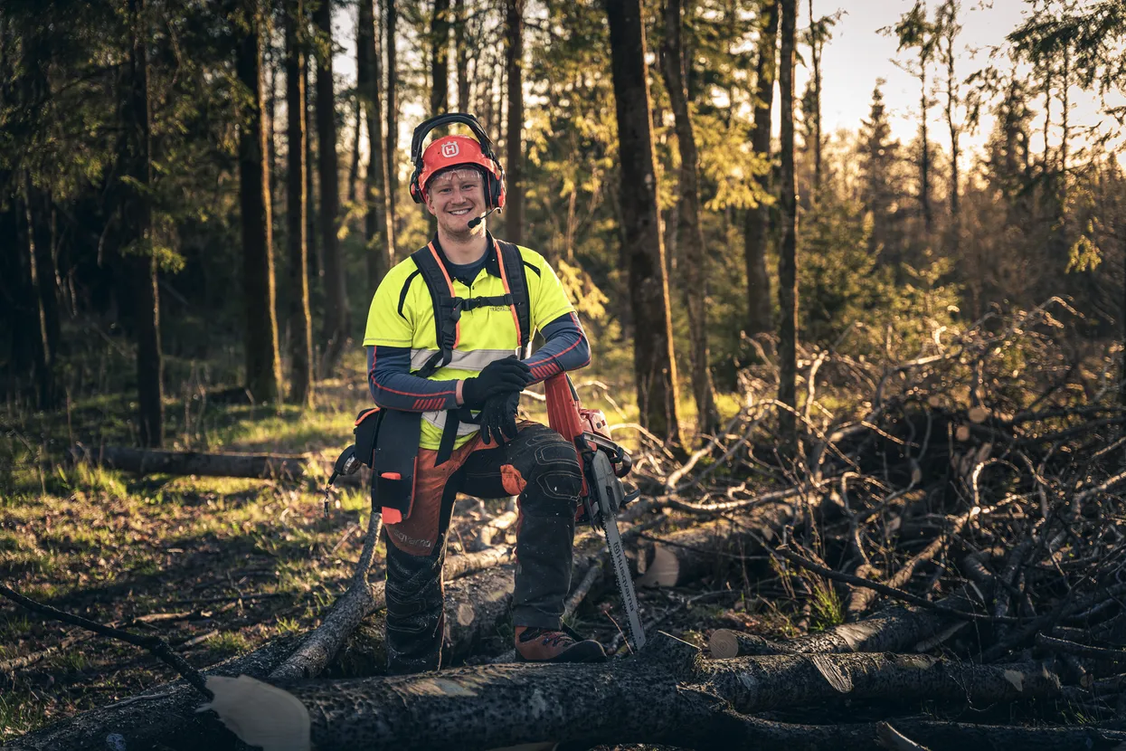 Smiling lumberjack wearing safety gear kneeling on a fallen tree in a forest with a chainsaw.