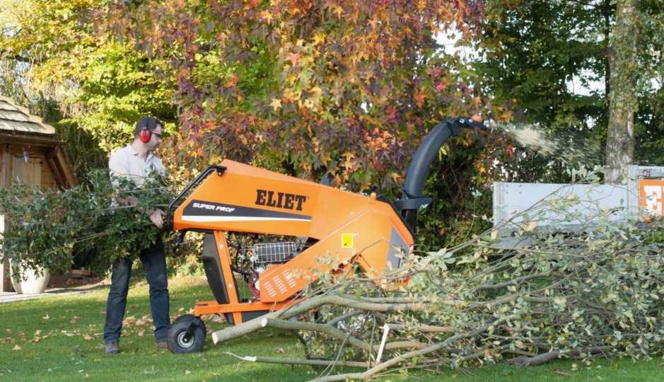 Man wearing ear protection operating an orange ELIET Super Prof wood chipper shredding tree branches outdoors on green grass.