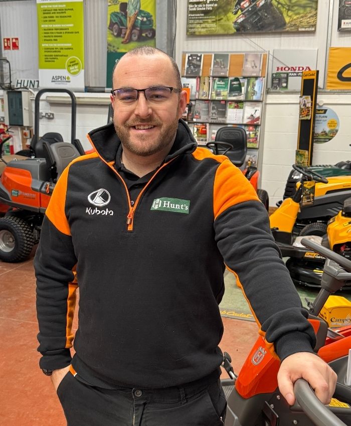 Smiling man wearing glasses and a black and orange Hunt's and Kubota branded jacket standing inside a store with lawn mowers in the background.