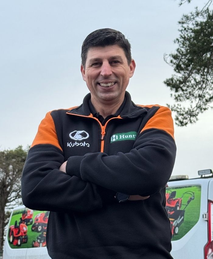 Smiling man with arms crossed wearing a Kubota and Hi Hunter branded jacket, standing outdoors near a van with images of lawn mowers.