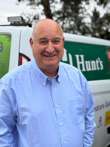 Smiling bald man wearing a light blue button-up shirt standing in front of a white van with a green and red Hunt's logo.