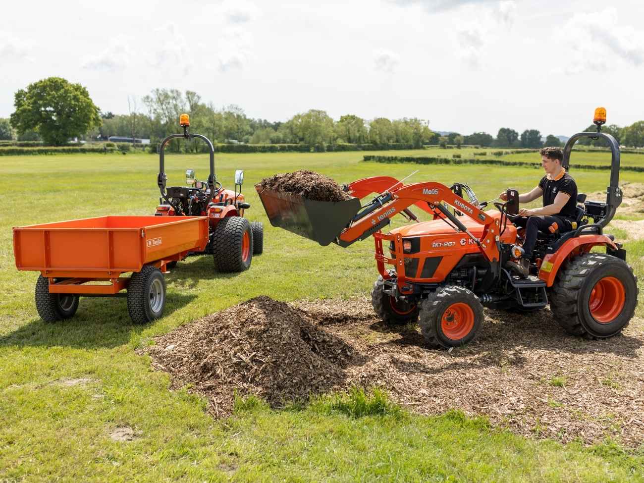 Man operating an orange Kubota tractor with loader to scoop mulch into a matching orange trailer in a green field.