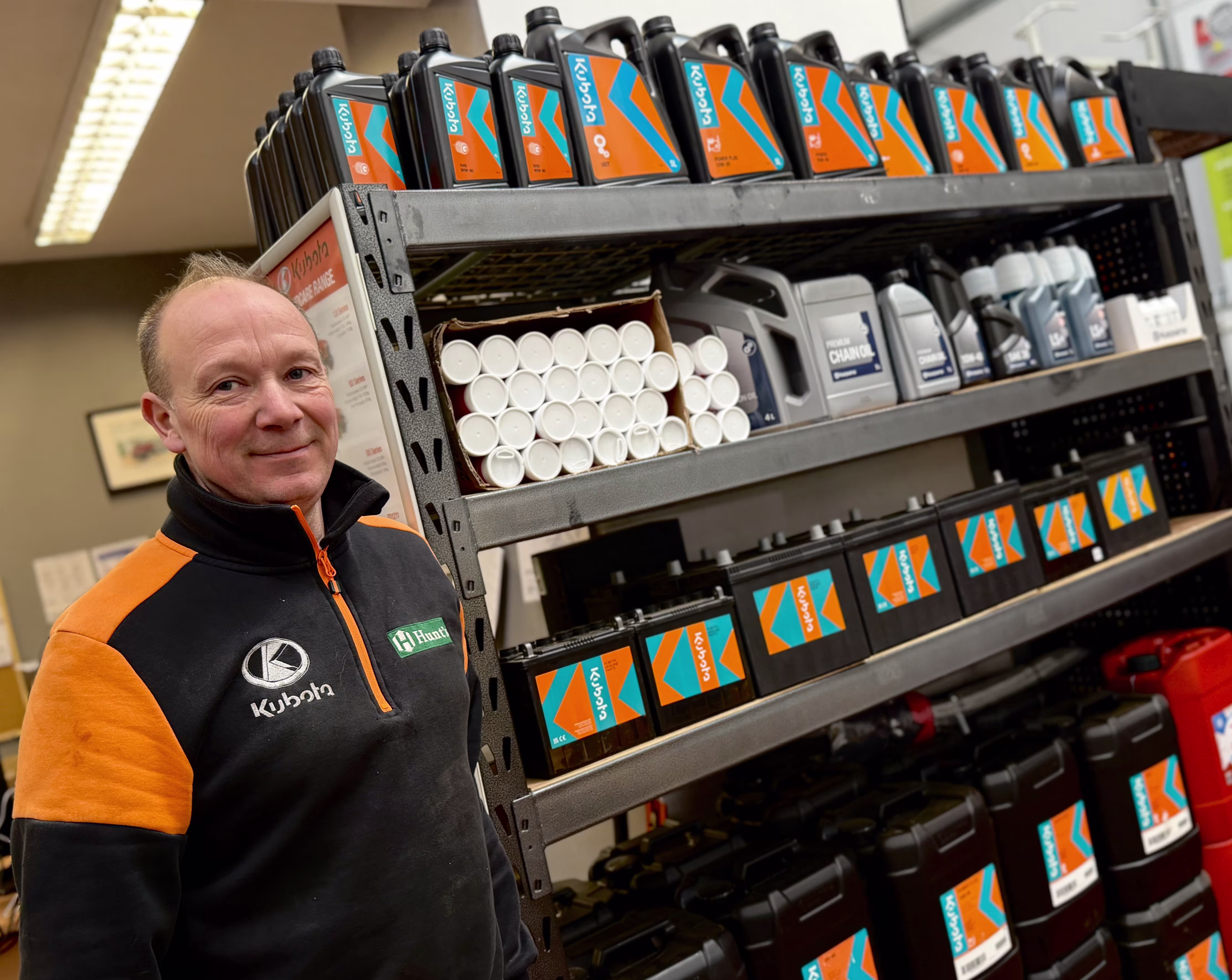Man in a black and orange Kubota jacket standing next to metal shelves stocked with Kubota-branded oil and lubricant containers.
