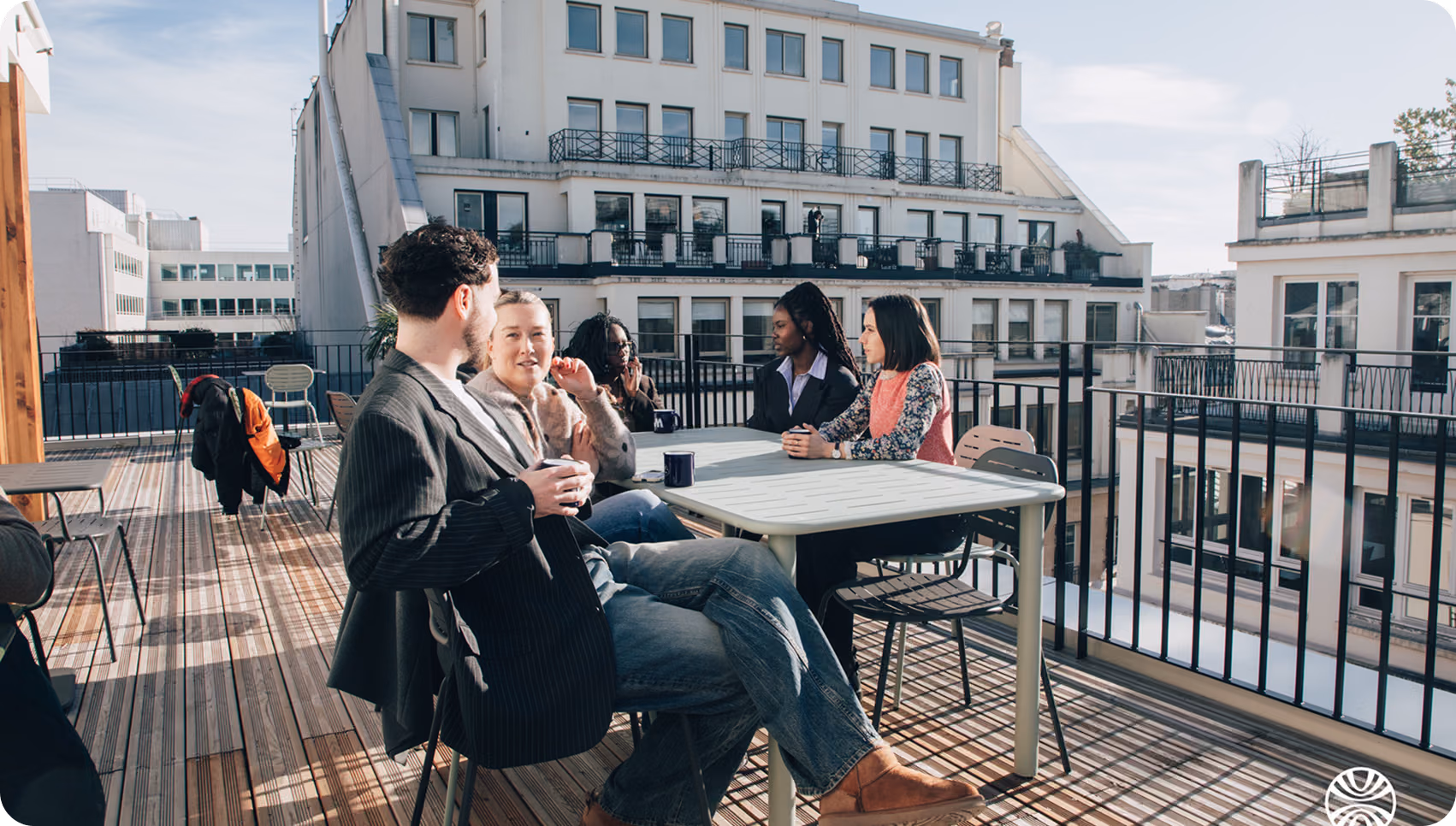 Groupe de personnes assises à une table sur une terrasse urbaine ensoleillée discutant.