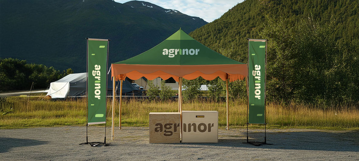 Grønn og oransje messe- eller salgsstand med teksten «agrinor» på teltet og to grønne bannere på hver side, plassert utendørs med fjell og trær i bakgrunnen.