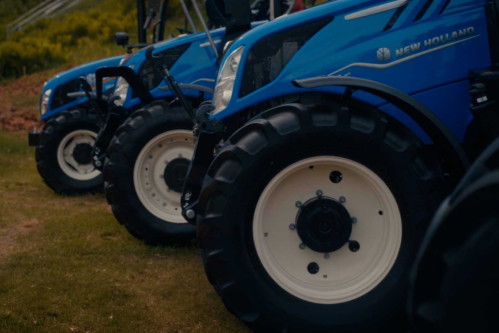 Nærbilde av hjul på en rekke blå New Holland-traktorer som står på gress.