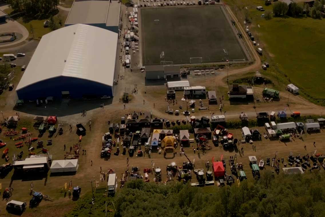 Luftfoto av et utendørs marked eller messe med flere boder, utstillinger av maskiner og kjøretøy, mennesker som går rundt, stort hvitt telt, og en fotballbane i bakgrunnen.