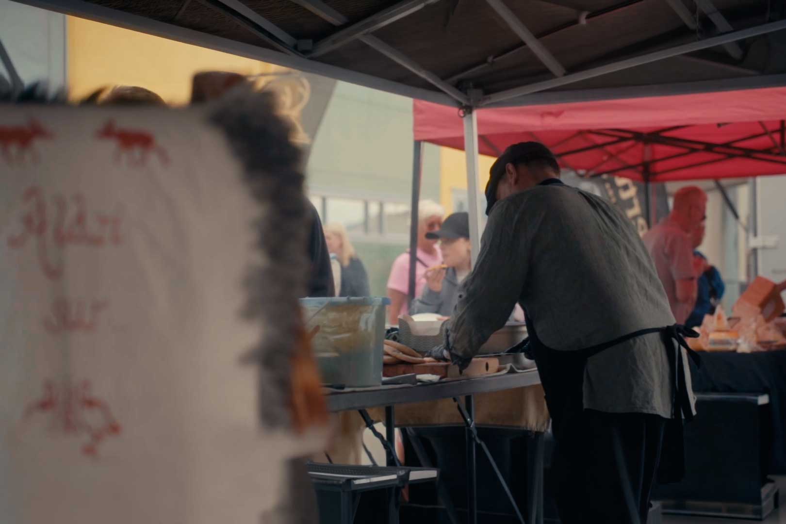 En mann i forkle og caps jobber ved et bord under et telt på et utendørsmarked, med folk som ser på eller spiser i bakgrunnen.