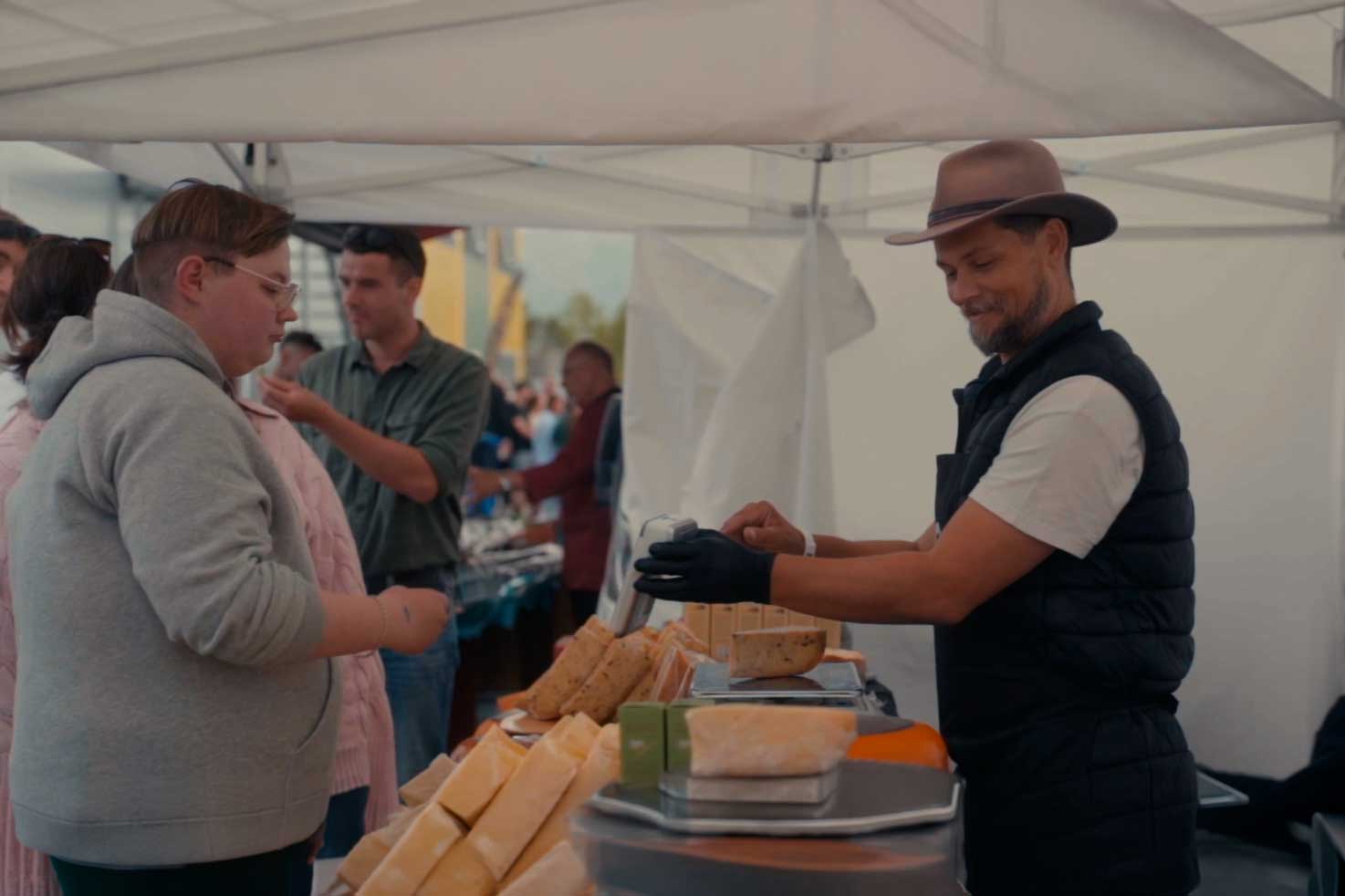 En mann med hatt og svart vest selger ost til en kunde i grå hettegenser under et markedstelt med folk i bakgrunnen.