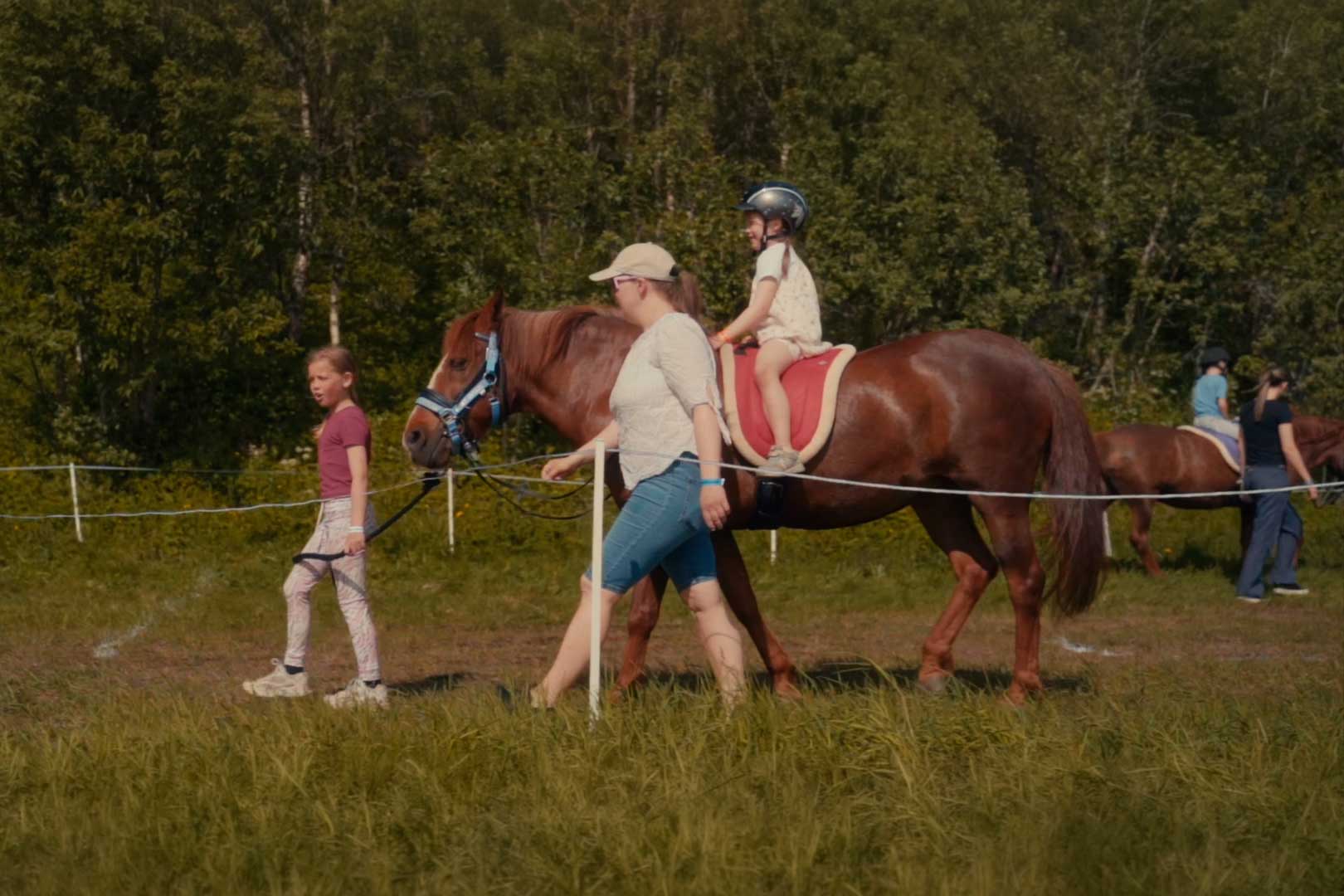 Barn som rir på en brun hest ledet av en voksen kvinne, med en jente som holder hestens tøyler foran dem, i et inngjerdet område med gress og trær.