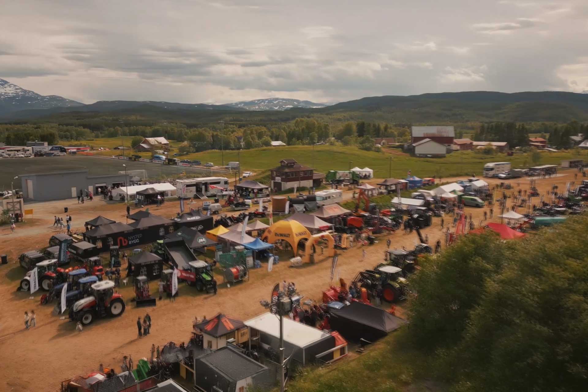 Oversiktsbilde av en utendørs messe med landbruksmaskiner som traktorer og utstillingsboder, omgitt av skog og fjell i bakgrunnen.
