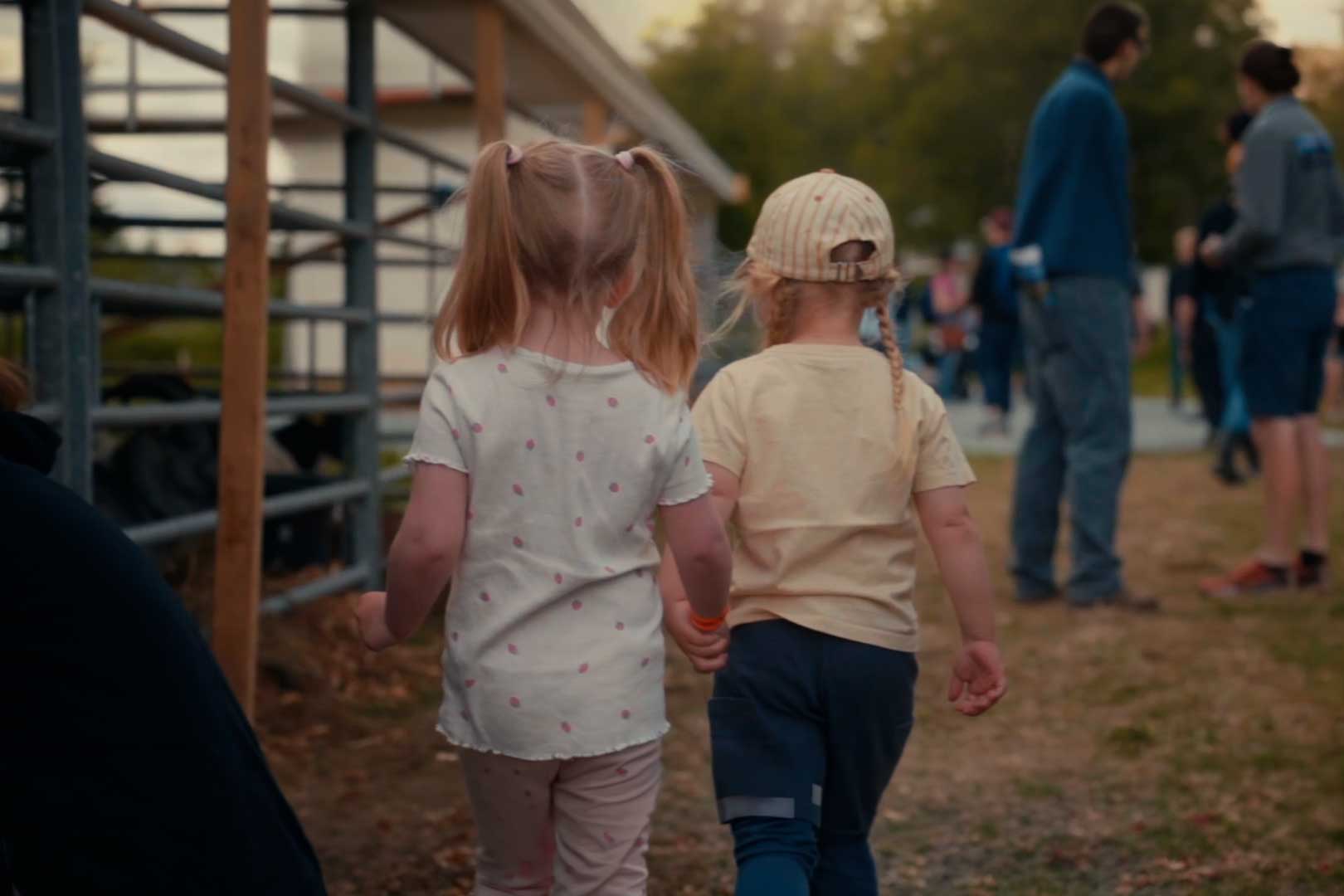 To små barn holder hender og går bortover en sti ute, med flere mennesker i bakgrunnen.