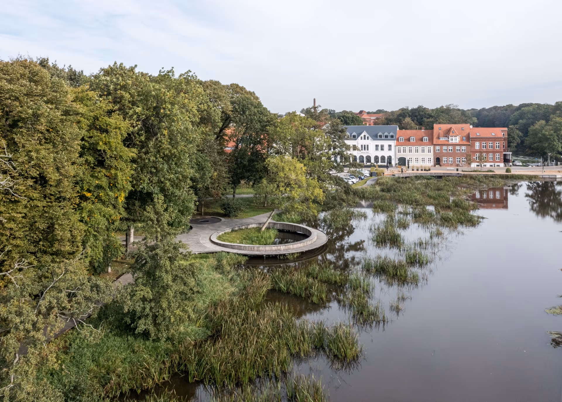 Vandringssti og cirkulær platform ved en sø med græs, træer, og historiske bygninger langs bredden.