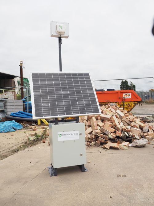Solar-powered environmental monitoring station with a solar panel, sensor unit, and rubble pile in the background.