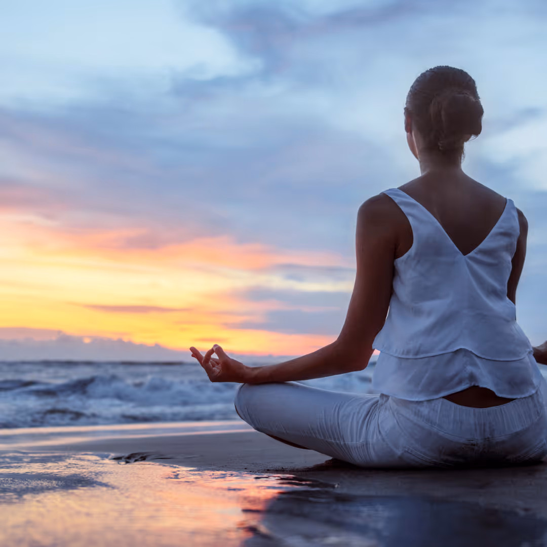 Person meditating in a seated yoga pose on the beach at sunset with colorful sky and ocean waves.
