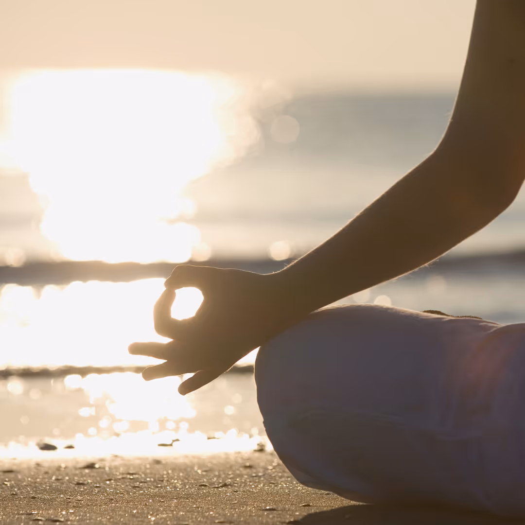 Person sitting cross-legged on the beach with hands in a meditation mudra during sunset.