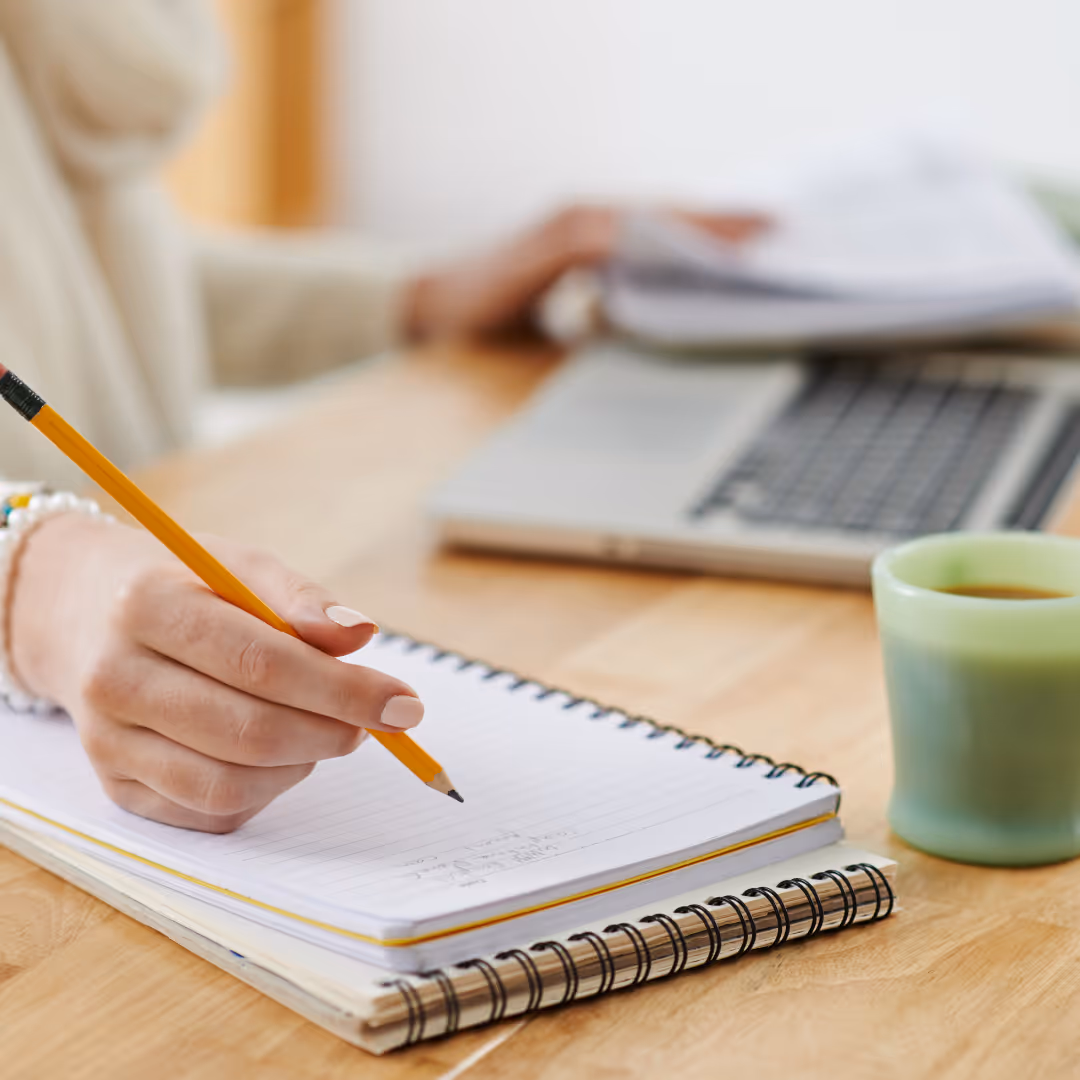 Person writing with a pencil on a spiral notebook beside a green cup and a laptop on a wooden table.
