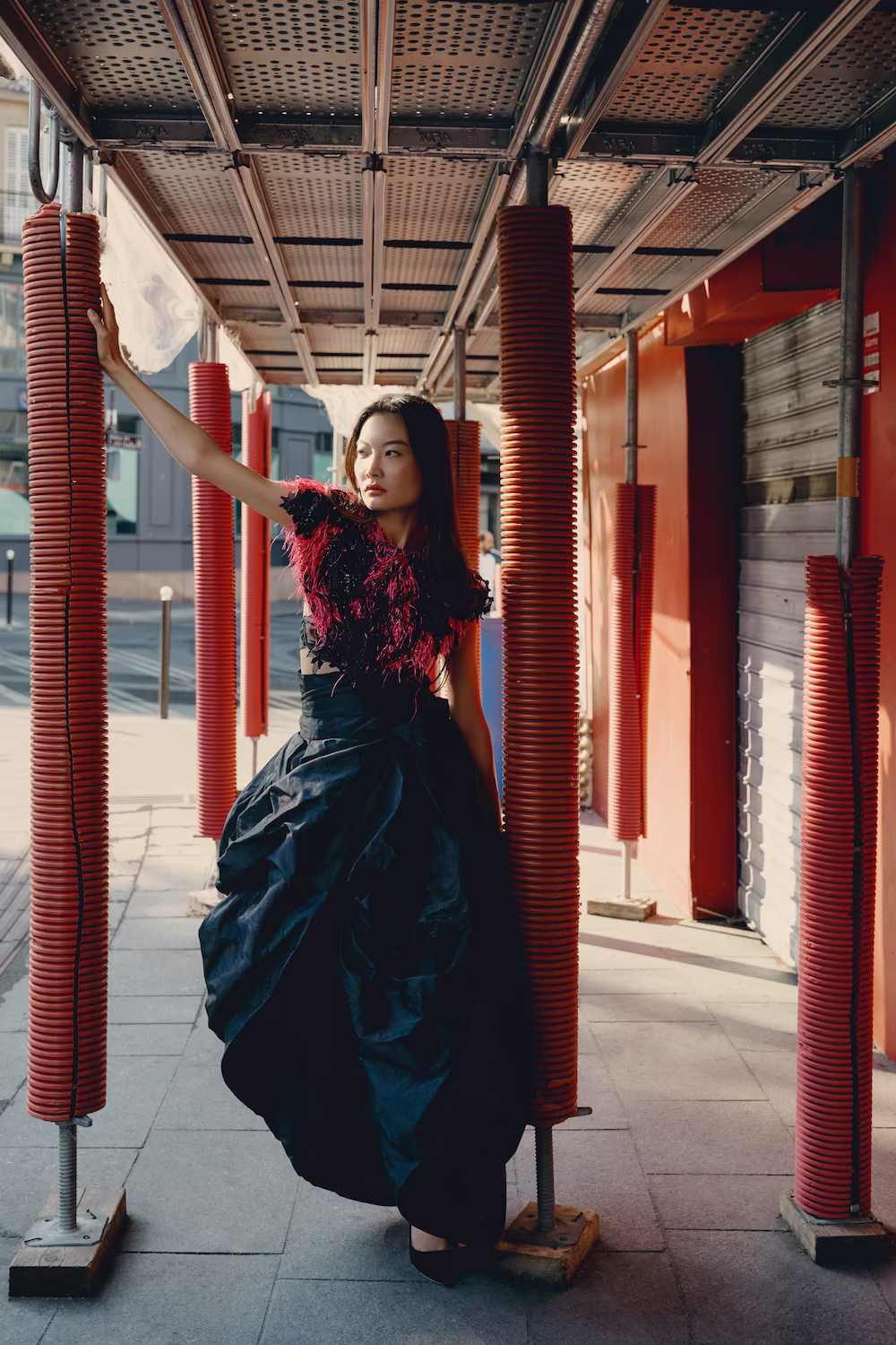 Model in a black feathered top and voluminous taffeta skirt in a red-columned Paris market arcade — fashion editorial