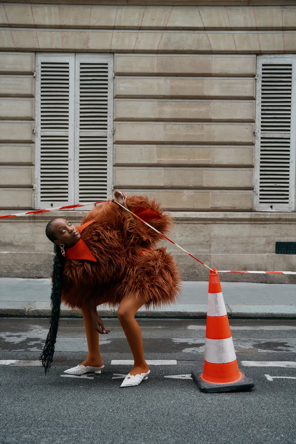 Model in a shaggy rust fur jacket bending beside a traffic cone on a Paris street — fashion editorial