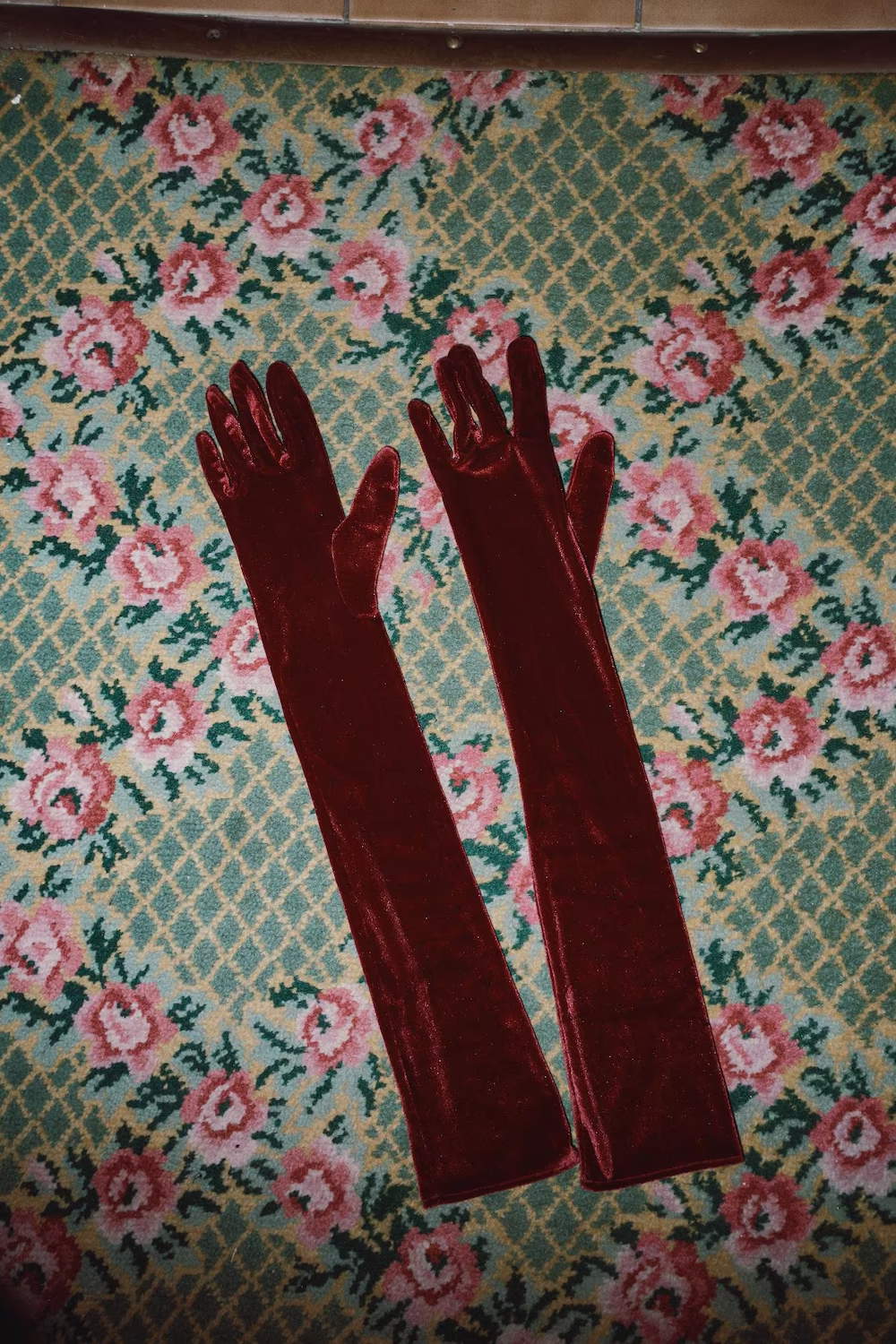 Still life of long burgundy velvet opera gloves on a vintage floral carpet — fashion editorial detail