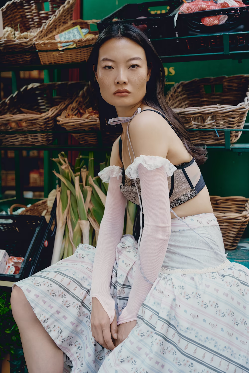 Model in a lace corset top and long gloves seated at a Paris greengrocer's stall — fashion editorial