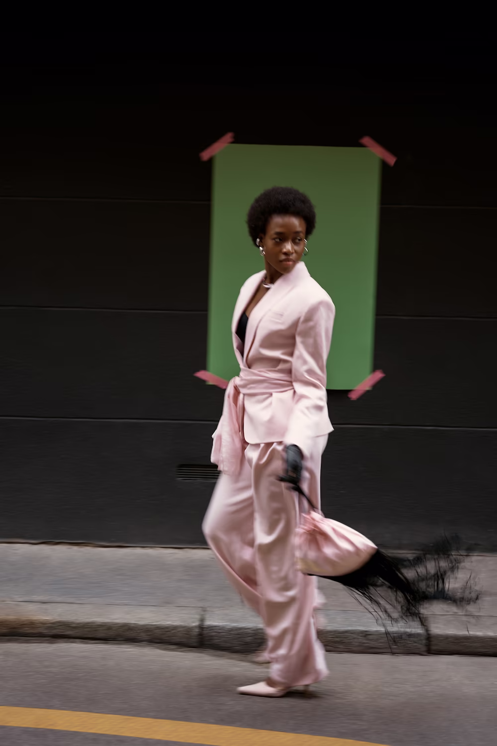Model in a pink satin suit walking past a green backdrop on a Paris street — fashion editorial