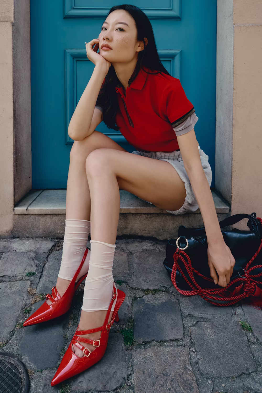 Model in a red shirt and denim shorts with red heels on cobblestones by a turquoise Paris door — fashion editorial