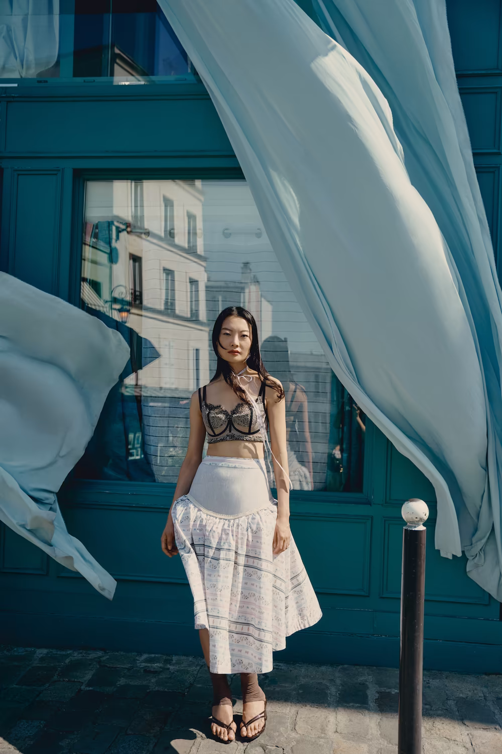 Model in a sheer embroidered top and printed skirt with fabric billowing outside a teal Paris shopfront — fashion editorial
